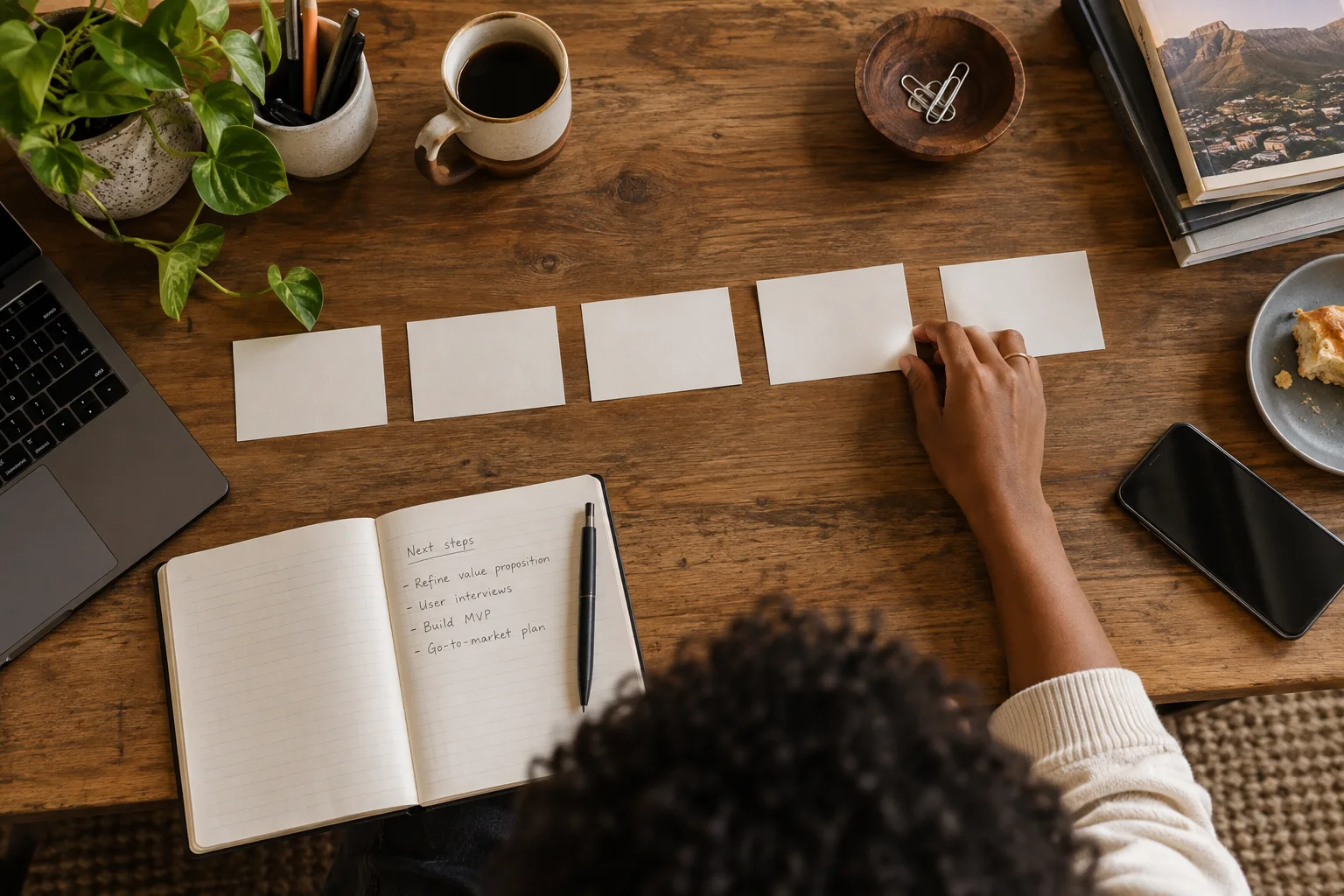 Top-down editorial photograph of a startup product planning desk: blank index cards in a neat sequence, open notebook, laptop edge and a single hand arranging priorities, warm neutral colours
